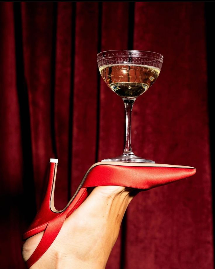 Red heel cradling a champagne coupe against velvet curtains